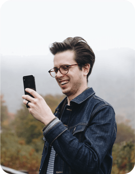 A young white man with facial hair and glasses looking at their phone whilst outside