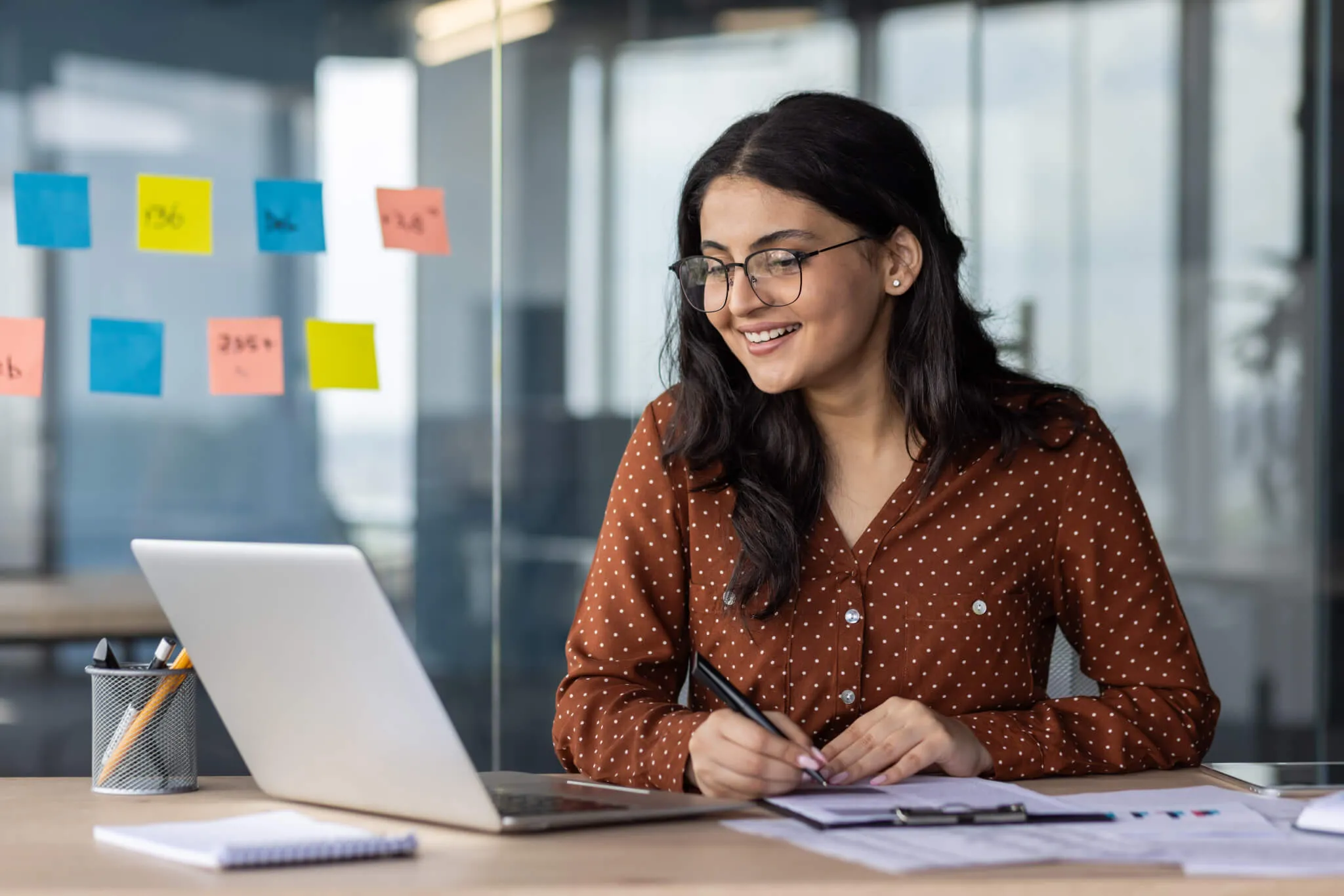 Happy woman financier doing paperwork inside office, latin american smiling pleased preparing financial report, accountant recording data and filling forms sitting at table with documents and laptop