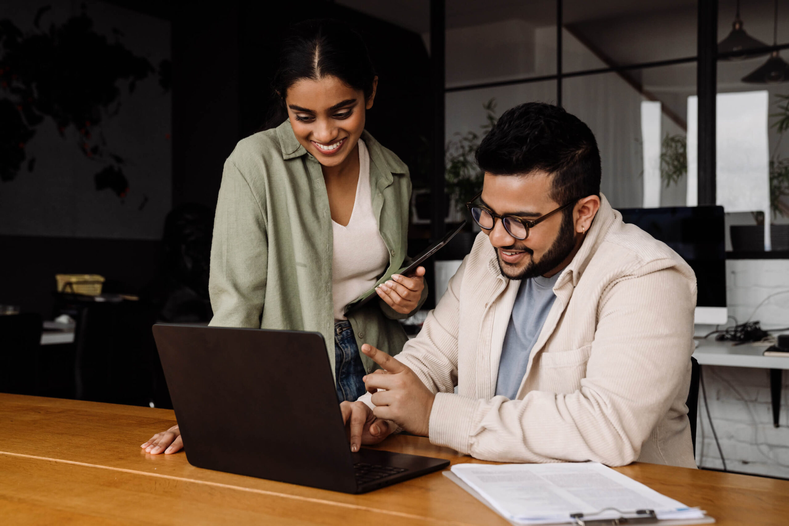 Two colleagues at desk collaborating over laptop, smiling and engaged in conversation.