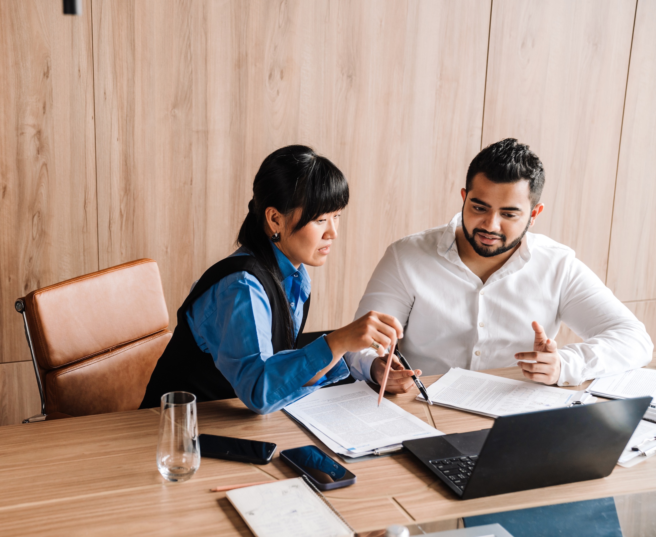 Two coworkers in modern office, reviewing documents and working on a laptop together.